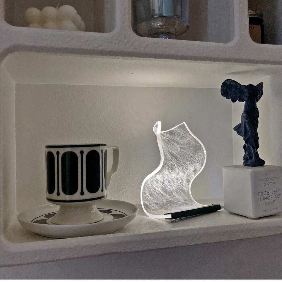 Warm high appearance level bedside lamp with abstract design emitting soft white light on shelf next to cup and sculpture