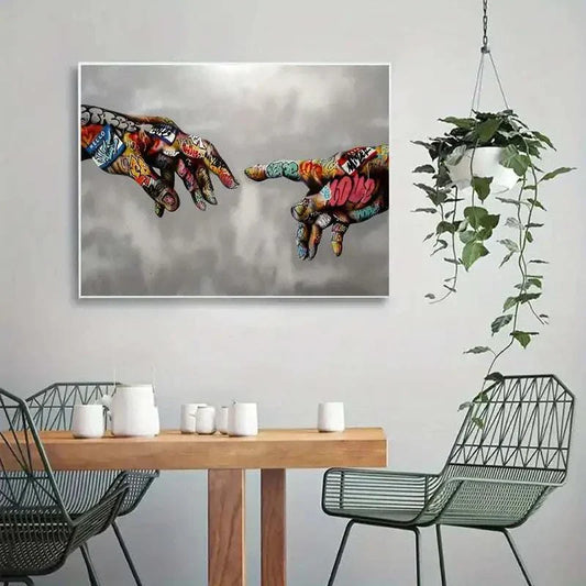 Canvas graffiti wall art featuring colorful spray-painted hands reaching towards each other, modern urban home decoration above a wooden dining table with metal chairs and a hanging plant.